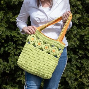Pu&ograve; includere: Una borsa a mano in crochet verde con bordi arancioni e un design a fiori in crochet. La borsa &egrave; tenuta da una persona che indossa una camicia bianca e jeans blu.