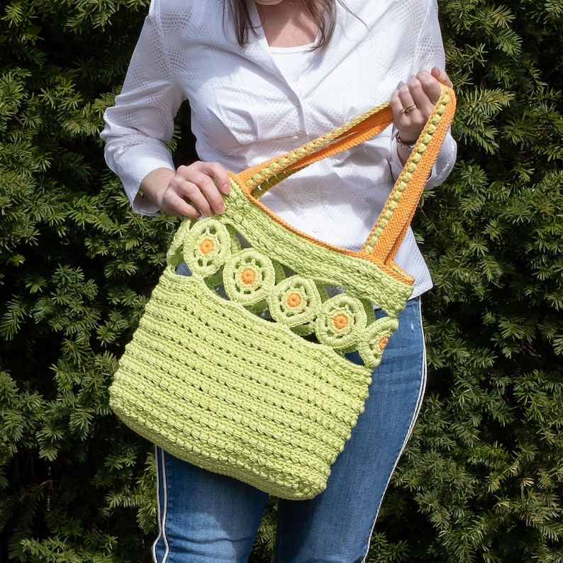 Pu&ograve; includere: Una borsa a mano in crochet verde con bordi arancioni e un design a fiori in crochet. La borsa &egrave; tenuta da una persona che indossa una camicia bianca e jeans blu.