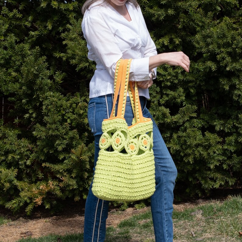 Pu&ograve; includere: Una borsa tote in crochet verde lime con bordi arancioni e un design floreale. La borsa ha una parte superiore larga e aperta e due manici lunghi.