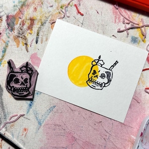 May include: A close-up of a yellow and black rubber stamp with a skull design. The stamp is on a white surface with a yellow circle around the skull.