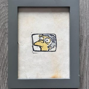 May include: A framed drawing of a yellow cartoon character with a large nose and gray hair. The character has a surprised expression with wide eyes. The drawing is on a white background and is framed in a dark gray frame.