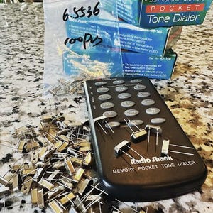 May include: A black Radio Shack Memory Pocket Tone Dialer with a pile of small gold-colored electronic components in front of it. The dial has a keypad with numbers, symbols, and the words "STORE", "DIAL", "PRS", "BTUV", "OPEN", "#", and "CANCEL". The dial also has the words "Radio Shack" and "MEMORY POCKET TONE DIALER" printed on it. There are three boxes stacked on top of each other in the background. The boxes are blue and white and have the words "33-Number Memory POCKET Tone Dialer" printed on them.