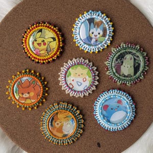 May include: Seven circular beaded pins showcasing Pokémon characters. Each pin features a colourful beaded border encircling a printed Pokémon image. The pins are displayed on a brown surface.