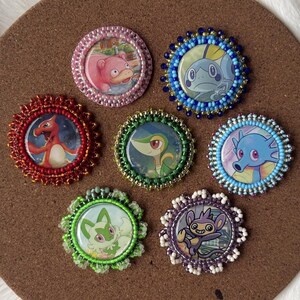 May include: Seven circular pins with cartoon characters, each with a beaded border. The characters are different on each pin, and the borders are in various colours, including red, green, blue, and purple. The pins are displayed on a brown surface.
