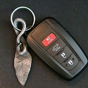 Pode incluir: Um porta-chaves de folha de metal feito à mão com um chaveiro de carro. O pingente em forma de folha tem detalhes gravados e é preso a um porta-chaves prateado. O chaveiro preto do carro tem botões vermelhos e brancos.