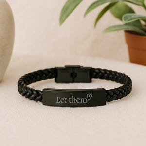 May include: A black braided leather bracelet featuring a matte black metal rectangular plate. The plate is engraved with the text "Let them" followed by a small heart symbol. The bracelet includes a matching black metal clasp closure.