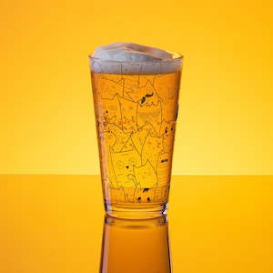 Op de afbeelding: Een hoog glazen bierglas met een wit schuimkopje. Het glas is versierd met een zwart-witte illustratie van katten.