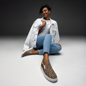 May include: A woman wearing a white denim jacket, blue jeans, and leopard print slip-on shoes. She is sitting on a gray background.