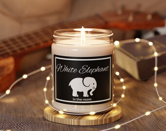 Könnte beinhalten: Eine weiße Kerze in einem Glasgefäß mit einem schwarzen Etikett, auf dem "White Elephant in the room" steht. Das Etikett zeigt auch eine weiße Silhouette eines Elefanten. Die Kerze ist angezündet und von weißen Lichterketten umgeben.