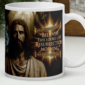 May include: White ceramic mug featuring an image of Jesus with the text "BELIEVER: THIS LOOKS LIKE RESURRECTION MORNING." The mug has a white handle and is set against a blurred background.