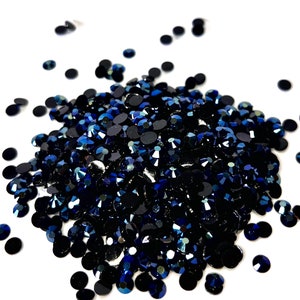 May include: A pile of dark blue iridescent rhinestones, scattered on a white background. The rhinestones are small and round, with a shiny, reflective surface. The text "Midnight Lover" is written below the pile of rhinestones.