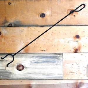 May include: Black metal fireplace poker with a hook on one end and a double prong on the other end. The poker is hanging on a wooden wall.