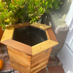 May include: A hexagonal wooden planter with a natural wood finish. The planter is made of multiple pieces of wood, creating a rustic look. The planter is empty and ready to be filled with plants.