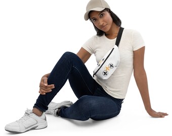 May include: A white fanny pack with black and gold fleur-de-lis designs. The fanny pack is worn by a person sitting on a white background.