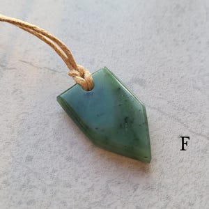 May include: A green jade pendant shaped like a pointed oval, hanging from a brown cord. The pendant has a mottled green colour and a smooth, polished surface.