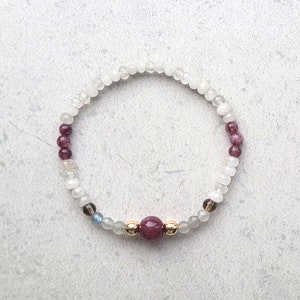 May include: A white and purple beaded bracelet with a single red gemstone in the center. The bracelet is made with small, round beads and has a gold clasp.