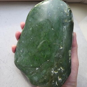 Peut inclure: Une grande pierre de jade verte polie, de forme lisse et arrondie. Le jade présente des variations de couleur, avec des zones vertes plus claires et plus foncées. La pierre est tenue dans une main, mettant en valeur sa taille et sa texture. Le jade est une pierre précieuse naturelle.