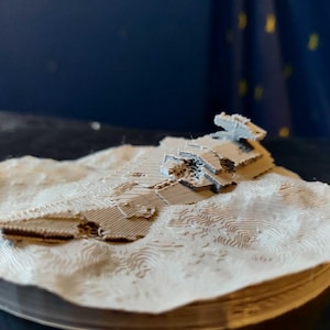 Puede incluir: Un modelo impreso en 3D de una nave espacial estrellada sobre una superficie texturizada. La nave espacial es gris y tiene un diseño detallado.