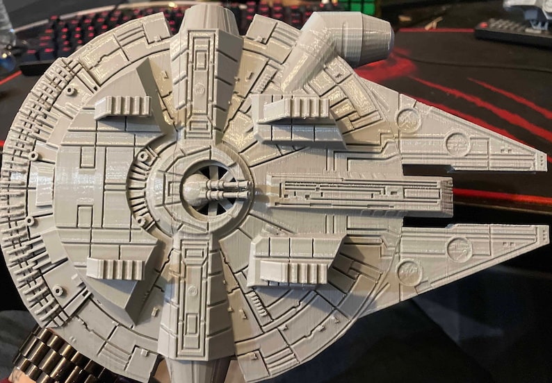 May include: A gray 3D printed model of the Millennium Falcon spaceship from Star Wars. The model is detailed with the iconic design features of the ship, including the cockpit, engine, and wings.