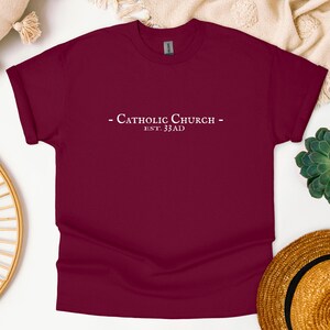 May include: Burgundy t-shirt with white text that reads "Catholic Church - Est. 33 AD"