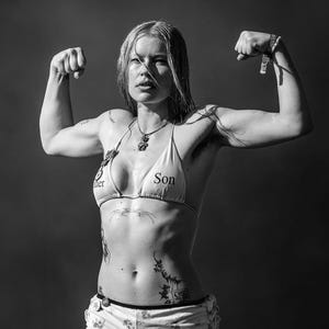 May include: Black and white image of a person flexing their arms. They are wearing a white bikini top with the word "Son" printed on it, and white shorts with a floral pattern and the number "03". The person has tattoos on their body.