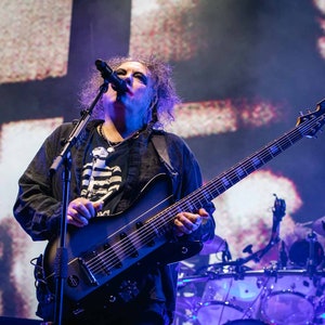 Puede incluir: Un músico con cabello oscuro y rizado canta en un micrófono mientras toca una guitarra eléctrica negra con un cuello largo y múltiples cuerdas. El fondo es una imagen borrosa de un escenario con una gran pantalla detrás del músico.