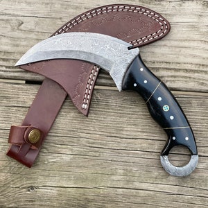 May include: A curved, silver-colored knife with a black handle and a leather sheath. The handle has a decorative pattern of white dots and a ring at the end.