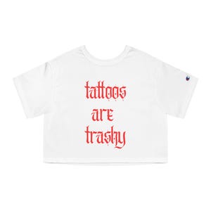 May include: White crop top with the text "tattoos are trashy" in red gothic font. The shirt has short sleeves and a Champion logo on the left sleeve.