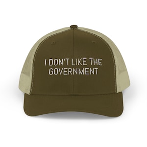 May include: An olive green trucker cap with a beige mesh back. The front of the cap has the white embroidered text, "I DON'T LIKE THE GOVERNMENT". The cap has a curved peak.
