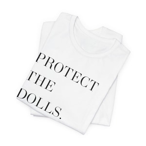 May include: White t-shirt with the text "PROTECT THE DOLLS." printed on the front.