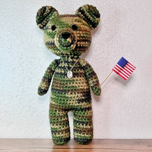 May include: A crocheted camouflage teddy bear holding a small American flag. The bear is wearing a silver necklace with a charm that says "Made with Love".