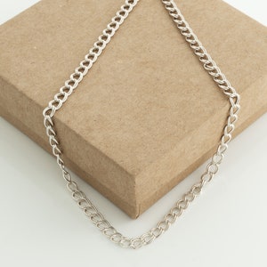 May include: A silver curb chain necklace displayed on a brown cardboard box. The chain is made of interlocking links, creating a continuous pattern. The necklace is a simple, elegant piece of jewelry.