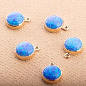 Könnte beinhalten: Fünf runde Anhänger aus blauem Opal-Edelstein mit goldfarbenen Fassungen und kleinen Schlaufen zur Schmuckherstellung. Die Opale haben eine lebendige, mehrfarbige blaue Farbe. Die Anhänger werden auf einer strukturierten, beigefarbenen Oberfläche präsentiert.