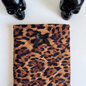 May include: A quilted tablet or e-reader case with a brown and black leopard print pattern. The case has a black bow at the top center. Two black skull-shaped planters with green plants are in the background.