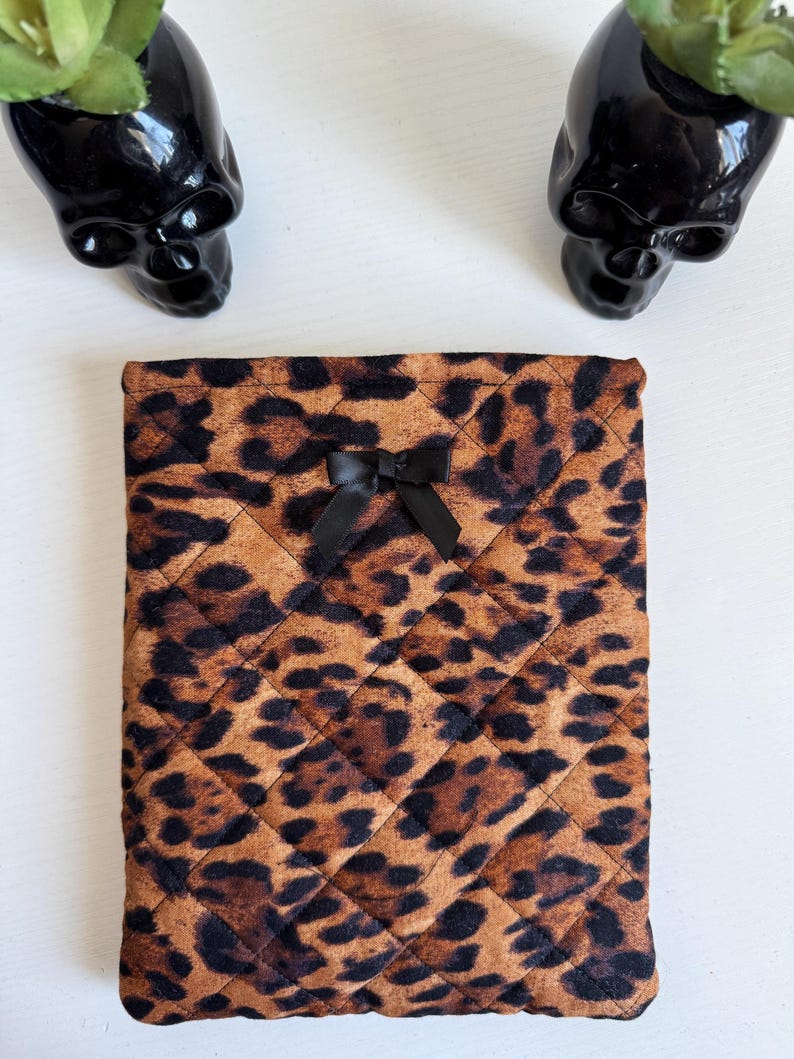 May include: A quilted tablet or e-reader case with a brown and black leopard print pattern. The case has a black bow at the top center. Two black skull-shaped planters with green plants are in the background.