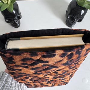 May include: A quilted book sleeve with a leopard print pattern, holding a book. The sleeve is brown and black, with a black interior. Two black skull planters with succulents are in the background.