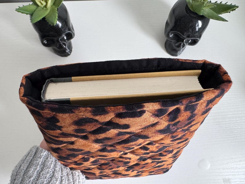 May include: A quilted book sleeve with a leopard print pattern, holding a book. The sleeve is brown and black, with a black interior. Two black skull planters with succulents are in the background.