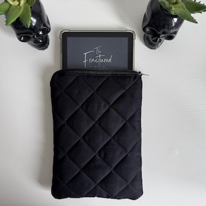May include: Black quilted tablet sleeve with a zipper closure, holding a tablet displaying the title "The Fractured". Two black skull planters with green plants are on either side of the tablet sleeve.