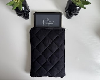 May include: Black quilted tablet sleeve with a zip closure, holding a tablet displaying the title "The Fractured". Two black skull planters with green plants are on either side of the tablet sleeve.