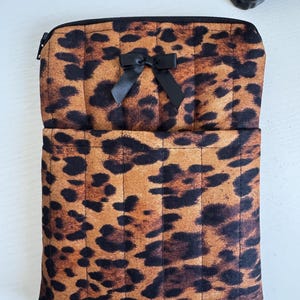 May include: A leopard print zippered pouch with a black bow. The pouch is brown with black spots and has a quilted design. The zipper is black. The pouch is open, revealing a pocket.