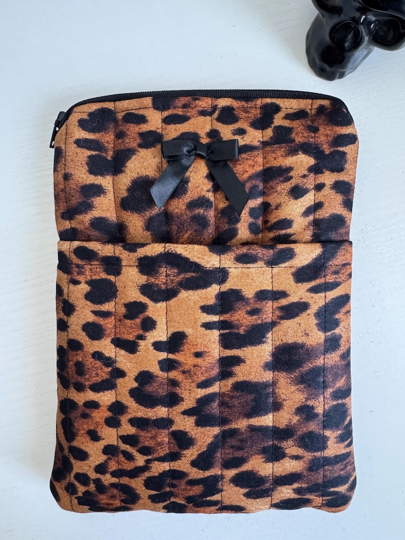 May include: A leopard print zippered pouch with a black bow. The pouch is brown with black spots and has a quilted design. The zipper is black. The pouch is open, revealing a pocket.
