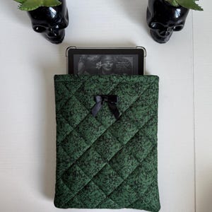 Könnte beinhalten: Eine gesteppte, waldgrüne Tablethülle mit schwarzem Blumenmuster und einer kleinen, mittig platzierten schwarzen Satinschleife auf der Vorderseite. Die Hülle enthält ein Tablet mit einem Bildschirm, der ein Gesicht und den Text Prepare to meet your next obsession zeigt.