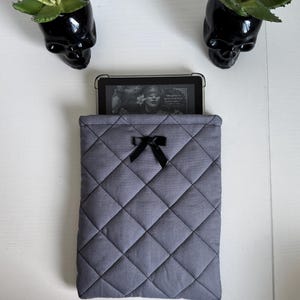 Könnte beinhalten: Eine graue, gesteppte Tablethülle mit einer schwarzen Schleife. Die Hülle hat ein Rautenmuster und enthält ein Tablet, das ein Buchcover anzeigt. Zwei schwarze, schädelartige Pflanzgefäße mit grünen Sukkulenten befinden sich auf beiden Seiten.
