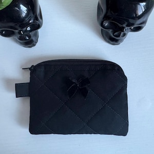 Puede incluir: Monedero negro acolchado con un pequeño lazo decorativo. El bolso tiene cierre de cremallera y una pequeña presilla de tela en el lateral. El fondo incluye dos macetas negras con forma de calavera.