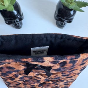 May include: A quilted cosmetic bag with a leopard print pattern, a black bow, and a black interior. The bag has a tag that reads "BOOKISH HEART". Two black skull-shaped planters with green plants are in the background.