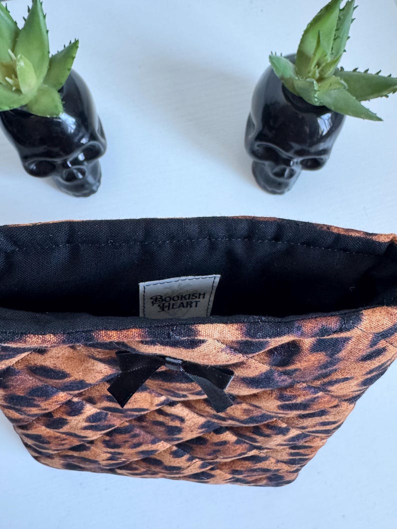 May include: A quilted cosmetic bag with a leopard print pattern, a black bow, and a black interior. The bag has a tag that reads "BOOKISH HEART". Two black skull-shaped planters with green plants are in the background.