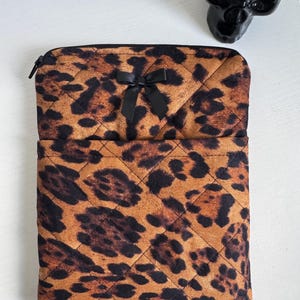 May include: A quilted leopard print case with a black zipper and a black bow. The case is a mix of brown and black spots. The case is rectangular with rounded corners.