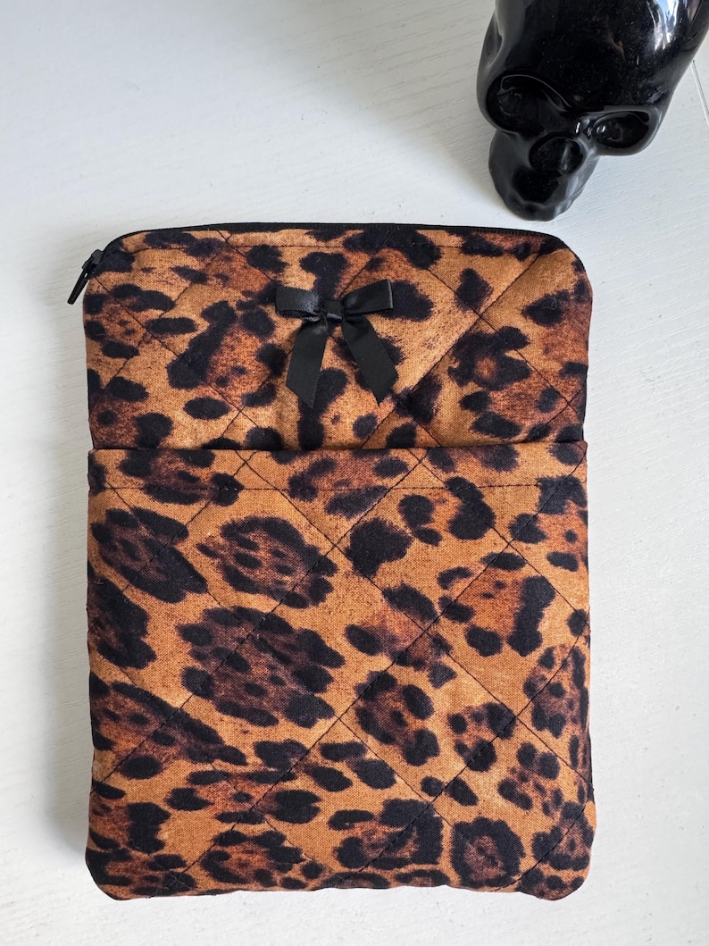 May include: A quilted leopard print case with a black zipper and a black bow. The case is a mix of brown and black spots. The case is rectangular with rounded corners.
