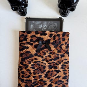 Puede incluir: Una funda acolchada para tableta con estampado de leopardo y un lazo negro. La funda contiene una tableta que muestra la portada de un libro. Dos macetas negras con forma de calavera con plantas verdes a cada lado.