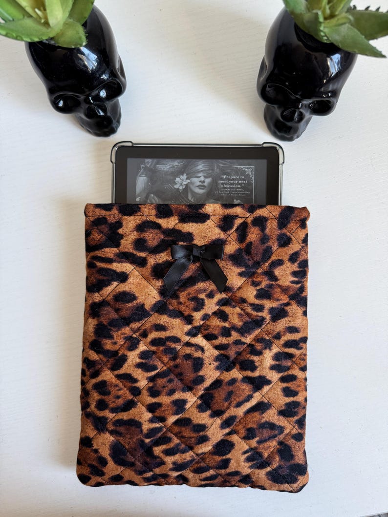 May include: A quilted leopard print tablet sleeve with a black bow. The sleeve holds a tablet displaying a book cover. Two black skull-shaped planters with green plants are on either side.